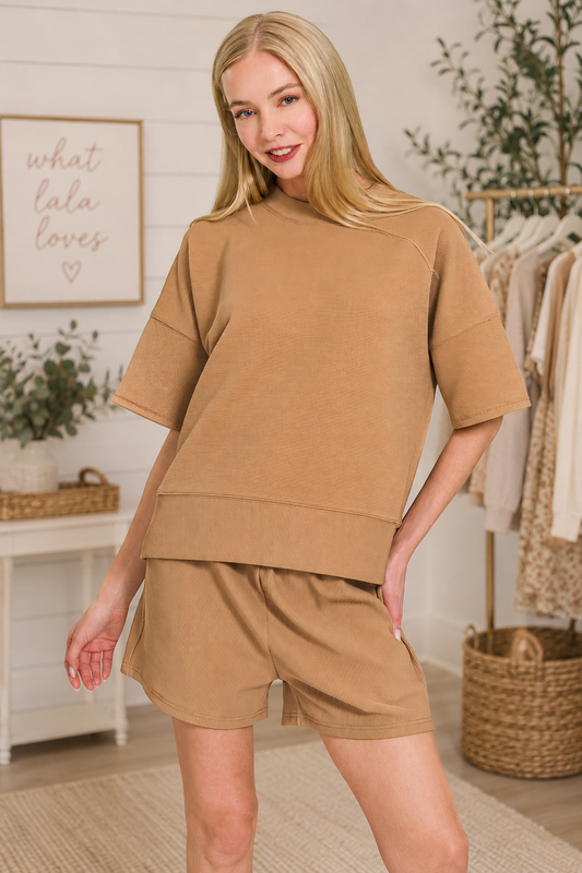 A woman modeling a matching tan lounge set consisting of a short-sleeve crewneck sweatshirt and coordinating shorts, set against a softly styled boutique background.