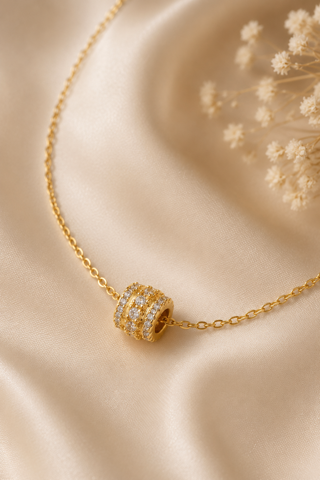Gold metal chain necklace with a barrel-shaped slider pendant featuring three rows of clear stones on draped fabric.