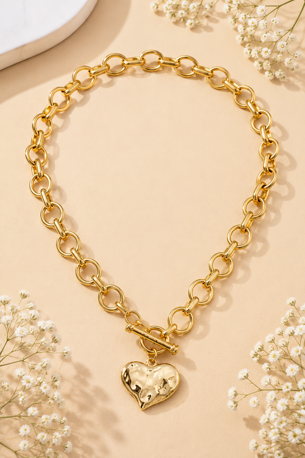 A gold chain necklace with a toggle clasp and a dangling heart-shaped charm, styled on a warm beige background with small white flowers.