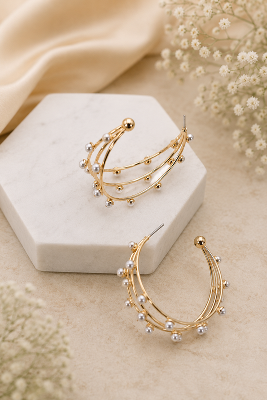 Gold crescent hoop earrings with delicate curved bands and small clear crystal-like beads, displayed on a white hexagonal block with cream fabric and baby's breath flowers.