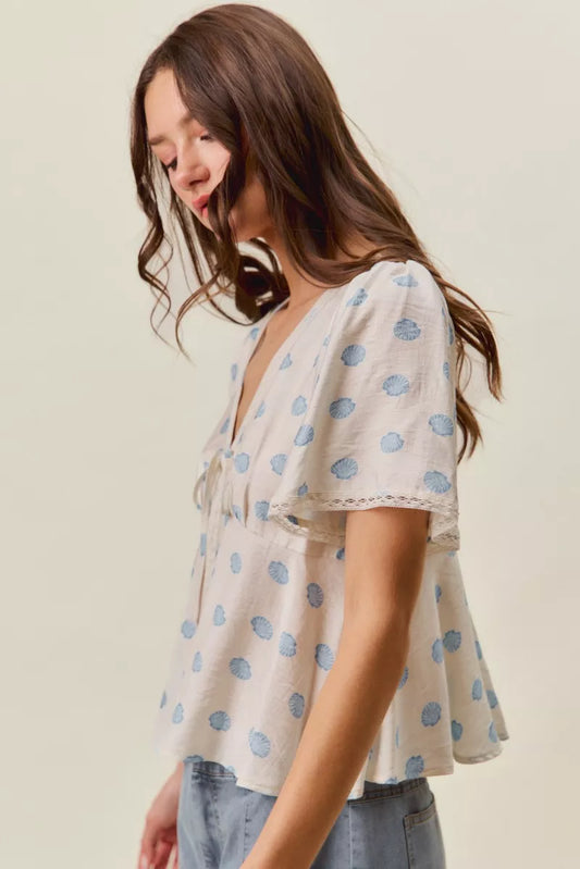 Woman wearing SO ME ivory blouse with blue shell print, tie-front neckline, flutter sleeves, and lace trim detail. Side View