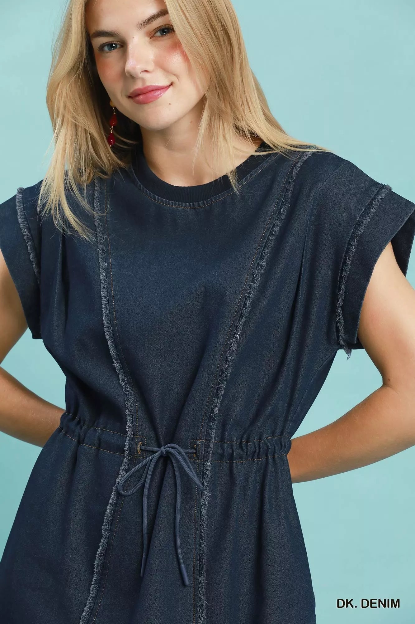 Woman wearing Umgee dark denim mini dress with raw edge seam detailing, drawstring waist, cap sleeves, and side pockets. Close View