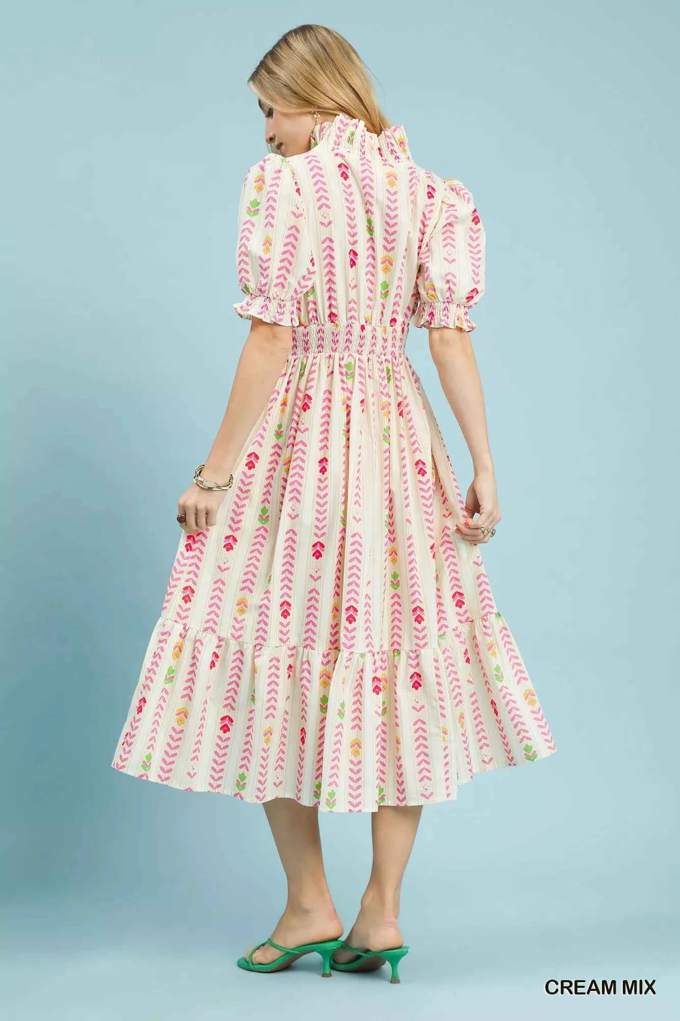 Woman wearing a cream floral print midi dress by Umgee featuring a smocked waist, tie neckline, puff sleeves, tiered skirt, and colorful spring-inspired pattern. back view
