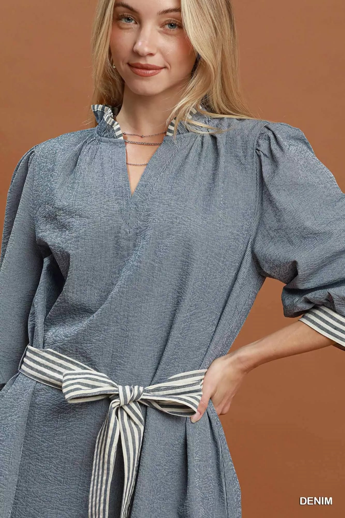 Woman wearing a denim blue textured mini dress with contrast striped trim, puff sleeves, side pockets, and a tie waist by Umgee. close view