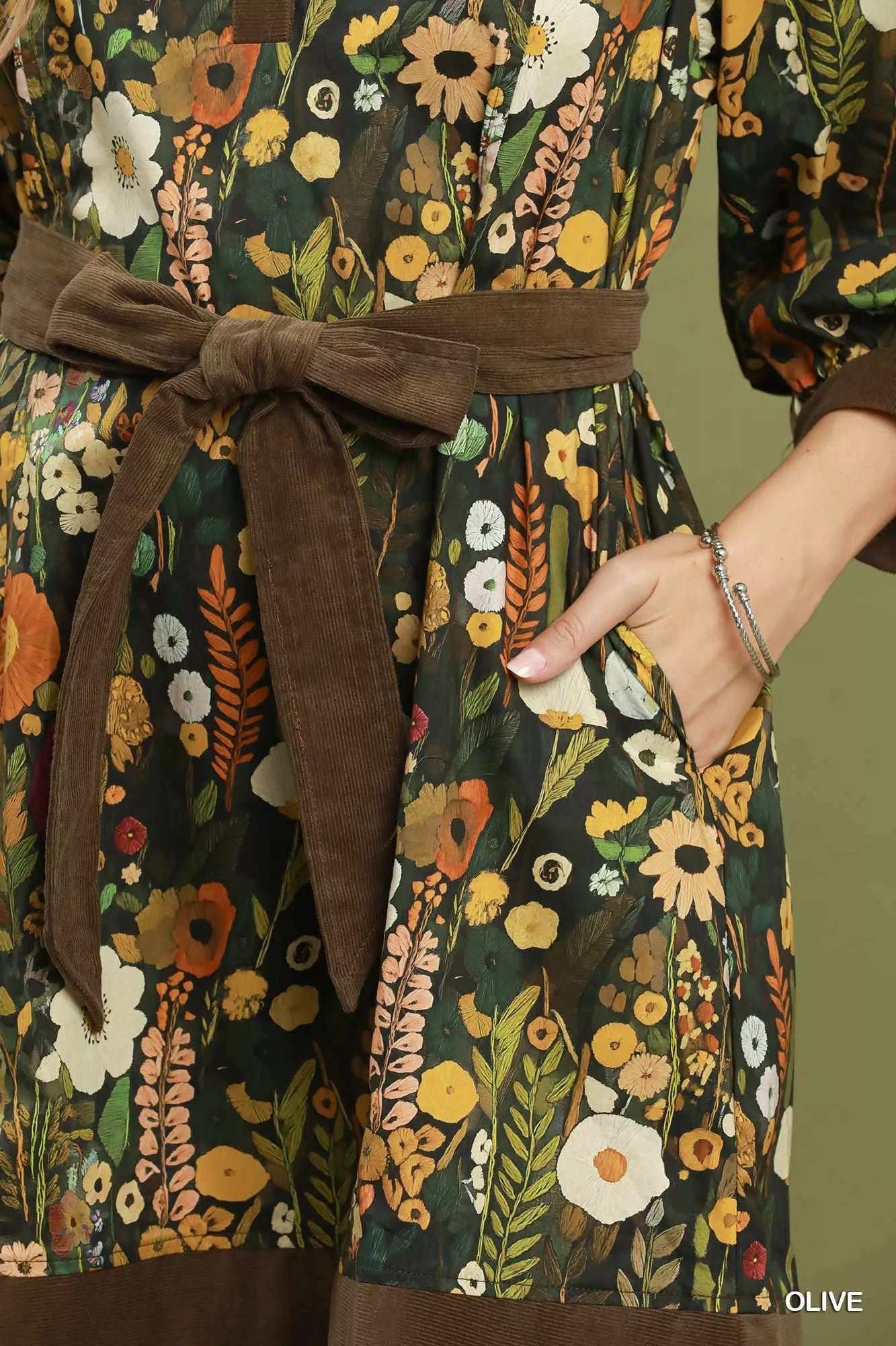 Woman wearing an olive floral print mini dress by Umgee featuring a collared neckline, puff sleeves, button-front detail, and a removable tie waist belt. pocket view