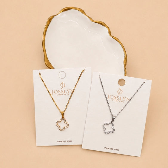 Two Josslyn Jewelry clover-shaped pendant necklaces on display cards, one gold-tone and one silver-tone, with a white dish with a gold rim behind them on a beige background.