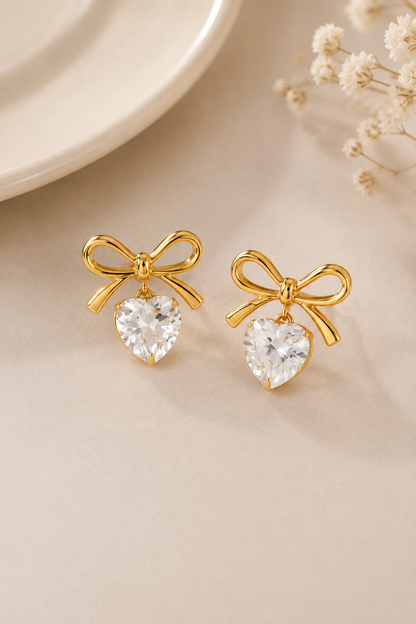 A pair of gold bow stud earrings with clear heart-shaped crystal drops, displayed on a light beige surface with a white plate edge and dried flowers in the background.