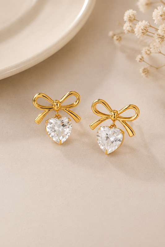 A pair of gold bow stud earrings with clear heart-shaped crystal drops, displayed on a light beige surface with a white plate edge and dried flowers in the background.