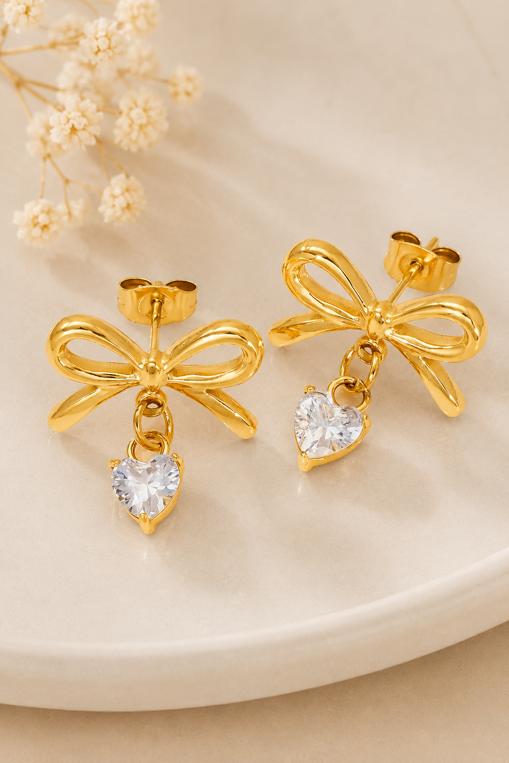 A pair of gold bow dangle earrings with clear heart-shaped crystal drops, displayed on a light beige ceramic plate with dried baby's breath flowers.