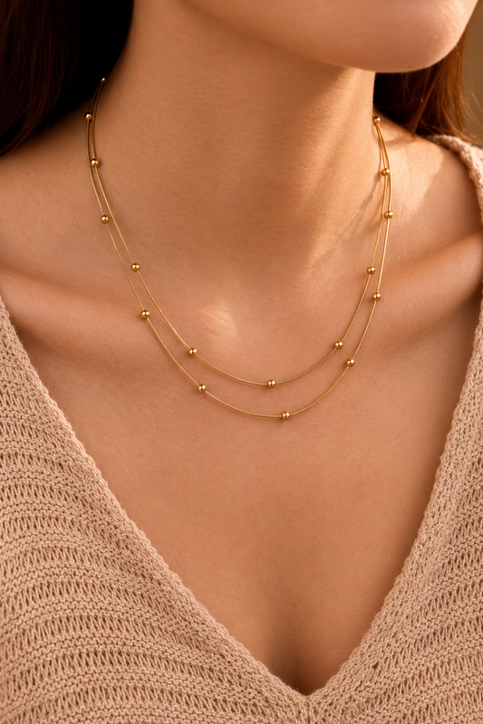 A close-up of a model wearing a delicate double-layered gold necklace with small bead details, paired with a beige knit V-neck sweater.