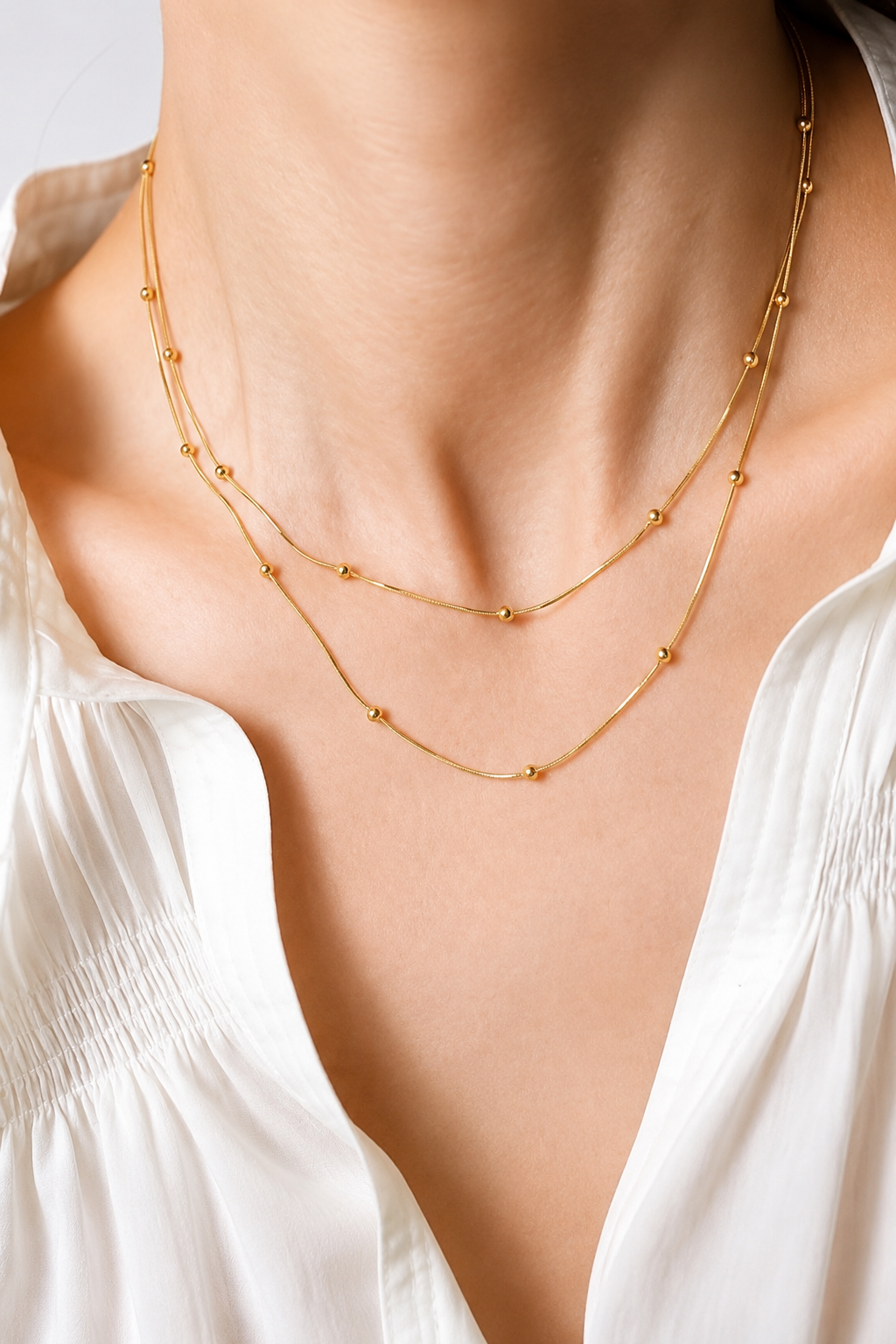 A close-up of a model wearing a delicate double-layered gold necklace with small bead details, paired with a white blouse.
