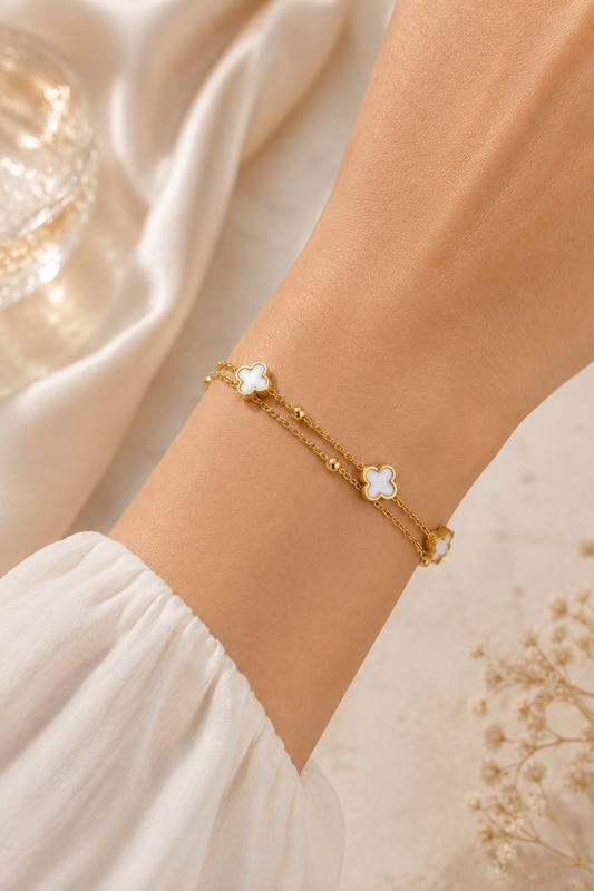 A delicate gold chain bracelet with three white clover-shaped charms, worn on a person's wrist. The background features soft white draped fabric and dried baby's breath flowers.