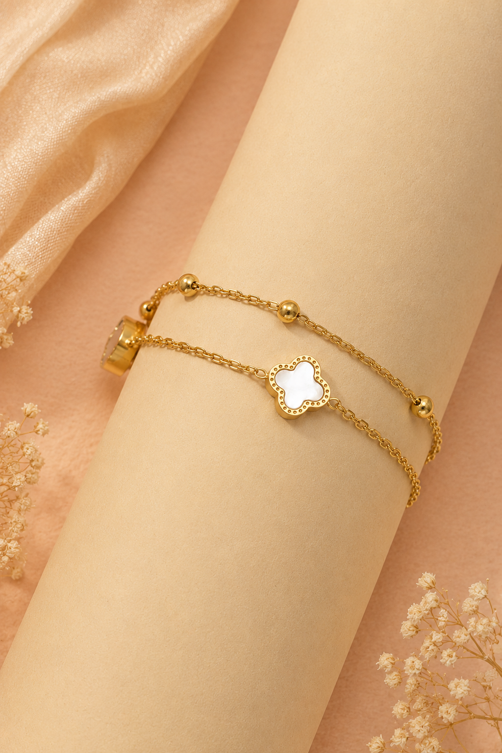 A delicate gold double-strand bracelet with a white mother-of-pearl four-leaf clover charm, displayed on a cream-colored wrist form against a soft peach background with draped satin and dried flowers.