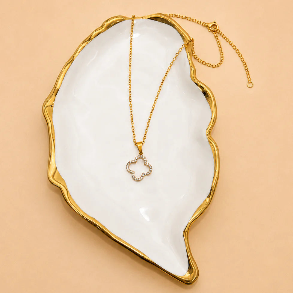 A gold chain necklace with a small flower-shaped pendant set with clear stones, displayed on a white and gold ceramic tray against a warm beige background.