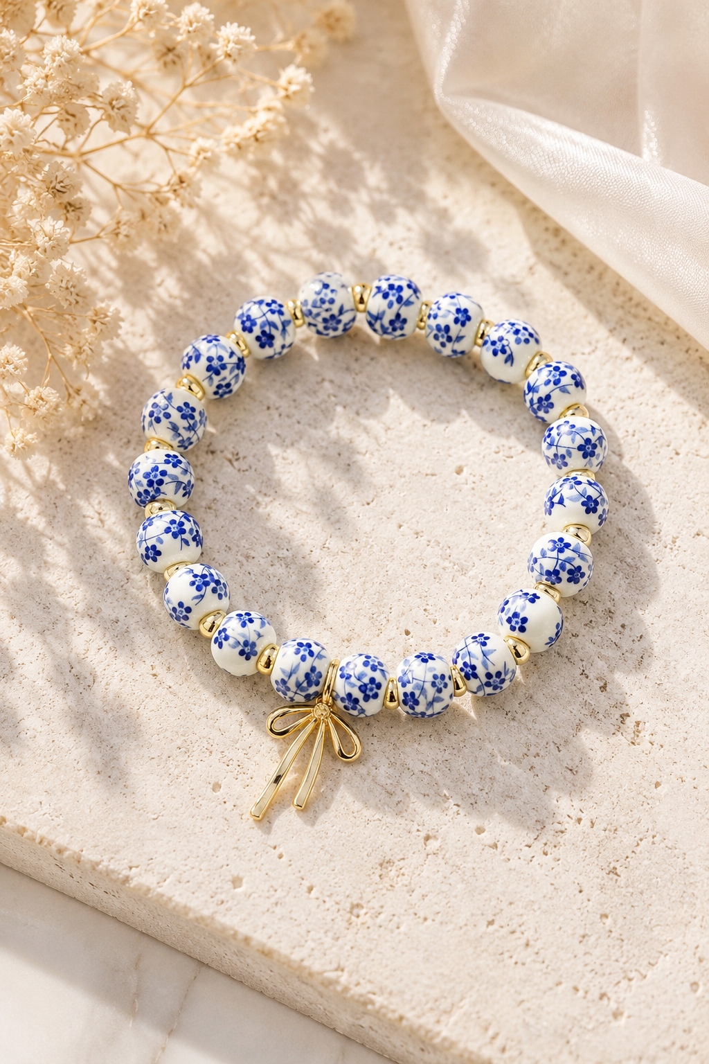 A beaded stretch bracelet with white and blue floral patterned beads, gold spacer beads, and a small gold bow charm, displayed on a textured beige stone surface with dried flowers and soft fabric in the background.