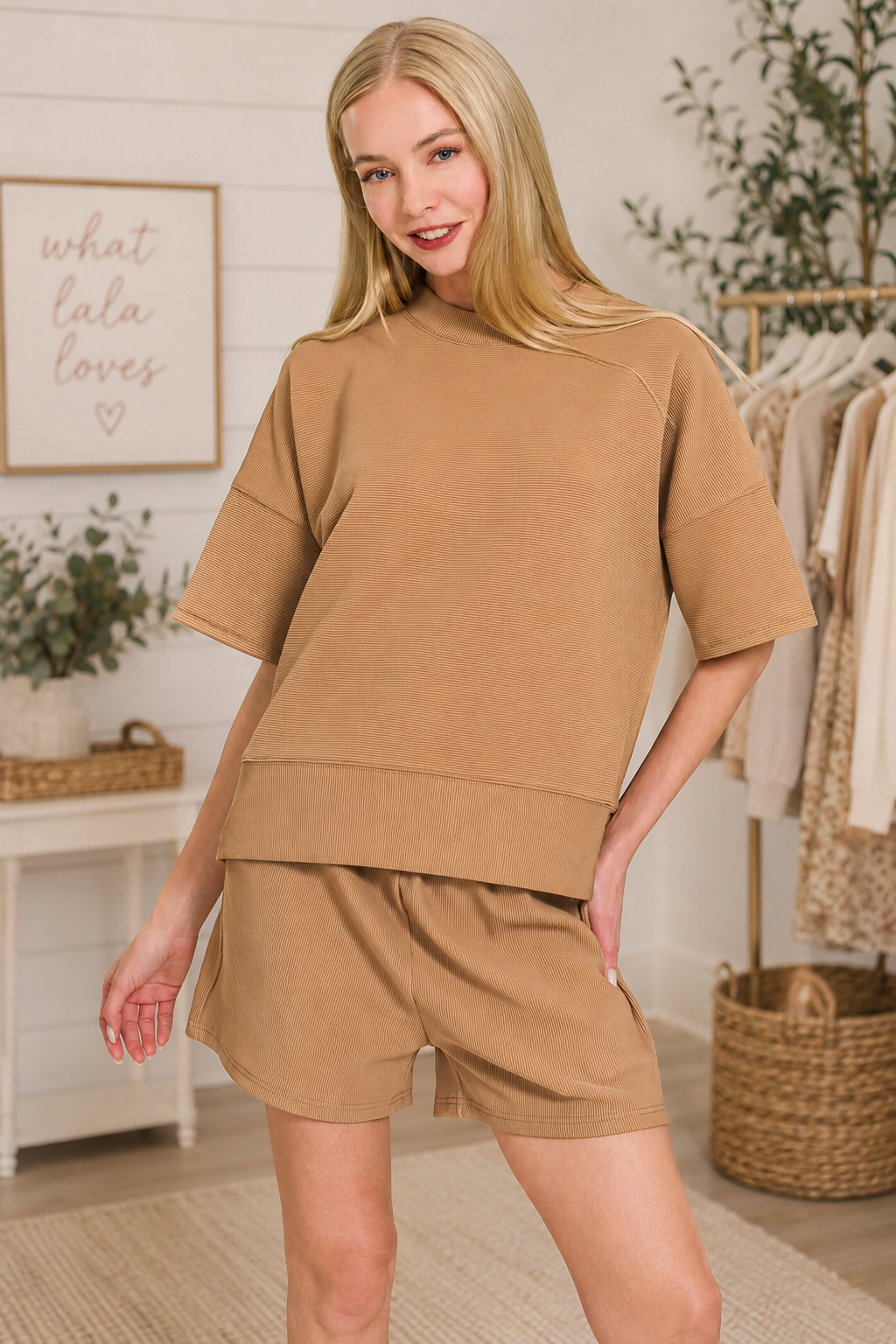 A woman modeling a matching tan lounge set consisting of a short-sleeve crewneck sweatshirt and coordinating shorts, set against a softly styled boutique background.