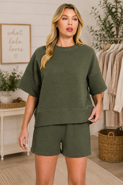 A woman modeling an olive green lounge set consisting of a short-sleeve crewneck sweatshirt and matching shorts, set against a cozy boutique background.