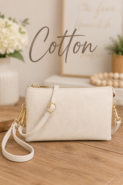 An off-white faux-cotton crossbody bag with gold-tone hardware, resting on a wooden table with a matching wristlet strap. The background features a warm, styled interior with a vase of white flowers, a wooden bead garland, and a framed print, with the word 'Cotton' written in script across the top.