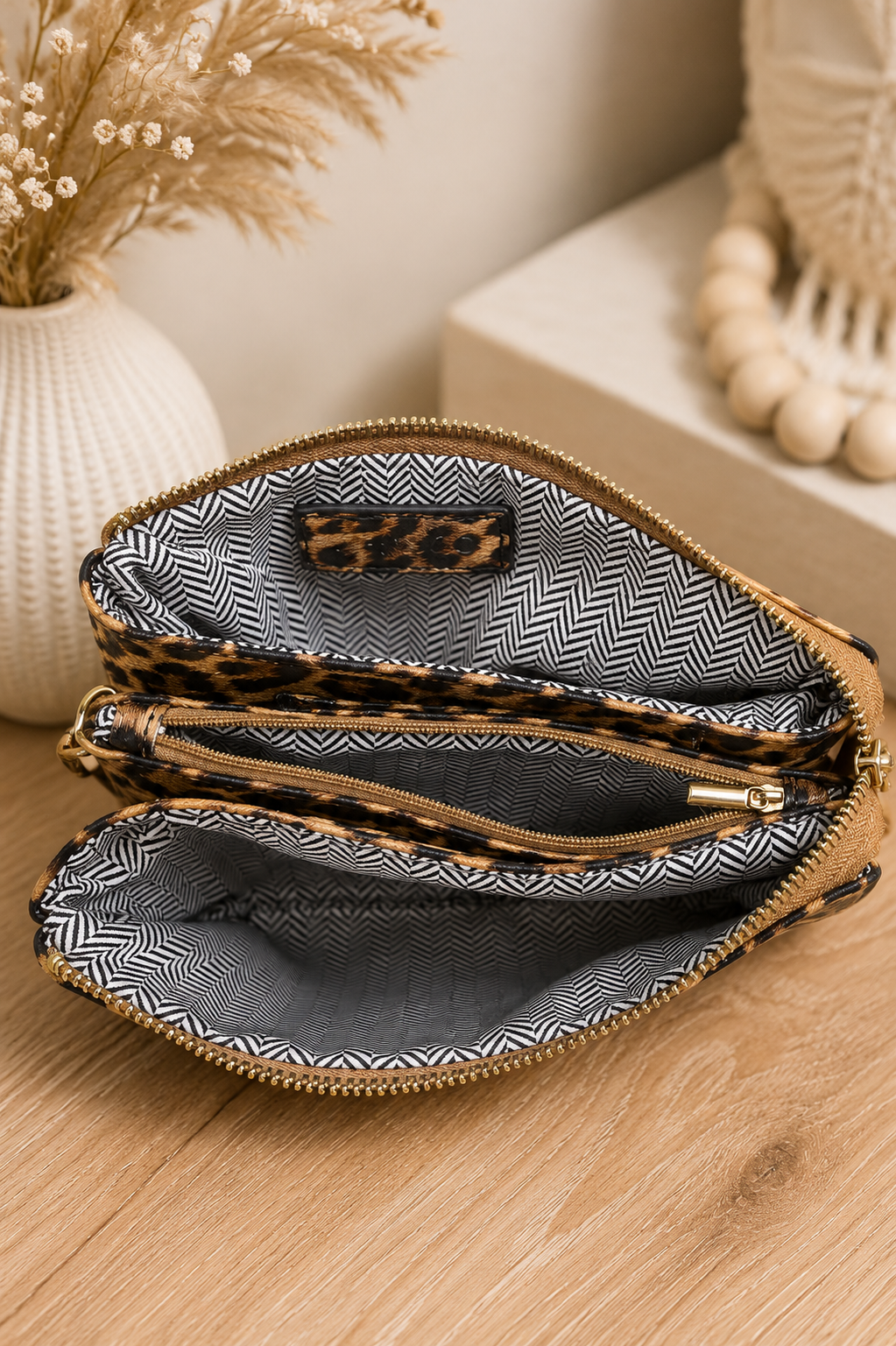 An open zippered clutch or pouch with a leopard-print exterior and a black-and-white herringbone-patterned interior, shown on a wooden surface with decorative home props in the background.