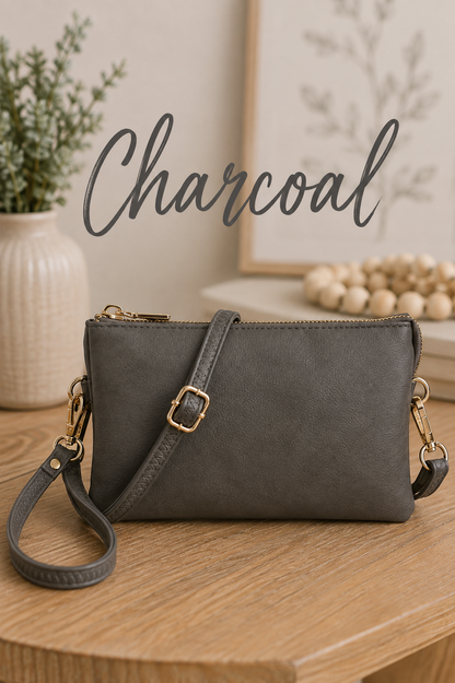 A charcoal gray faux-leather crossbody bag with gold-tone hardware, resting on a wooden table with a matching gray strap. The background features a neutral-toned room setting with a vase of greenery and a framed print, with the word 'Charcoal' written in script across the top.