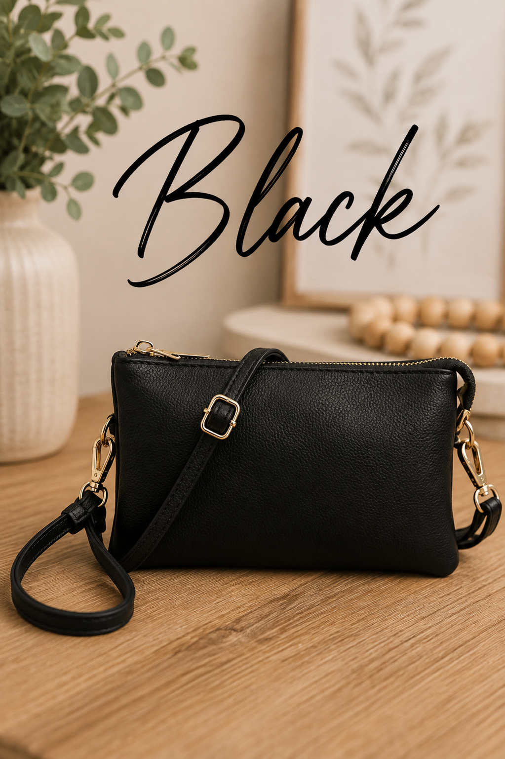A black pebbled faux-leather crossbody clutch bag with gold-tone hardware, shown on a wooden tabletop with a potted plant and framed art in the background. The word 'Black' is written in a black script font across the top.