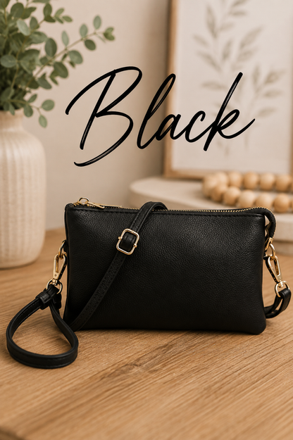 A black pebbled faux-leather crossbody clutch bag with gold-tone hardware, shown on a wooden tabletop with a potted plant and framed art in the background. The word 'Black' is written in a black script font across the top.