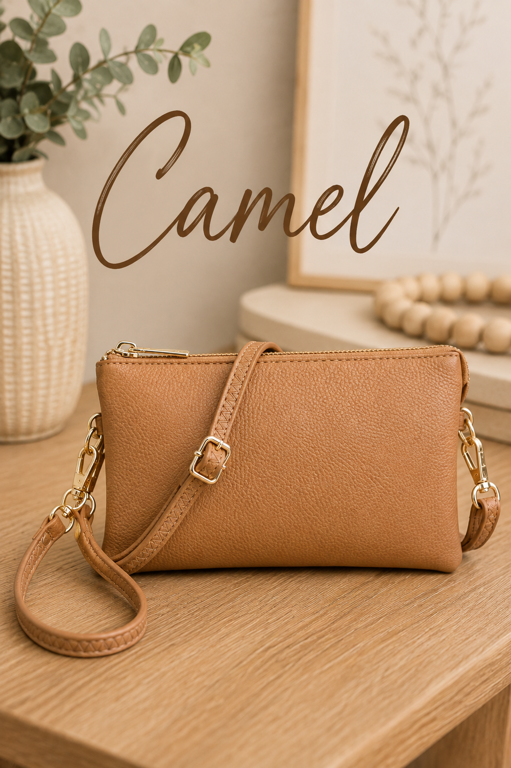 A camel tan faux-leather crossbody bag with gold-tone hardware, resting on a wooden table with a decorative background and the word 'Camel' written in script.