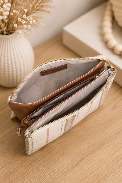 A beige and white textured zippered pouch with brown leather trim, shown open on a wooden tabletop with a neutral background and decorative props.