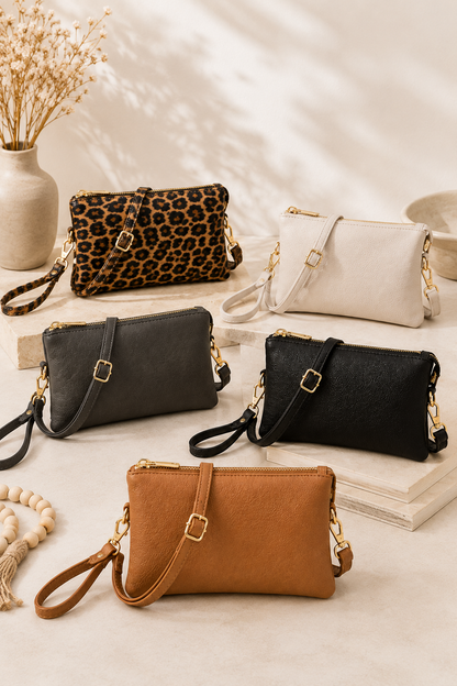 Five mini shoulder bags in various colors and textures are arranged on a beige surface with decorative props including a vase of dried flowers, books, and a beaded bracelet. The bags include leopard print, off-white, dark gray, black, and tan/cognac, each with a wristlet strap and gold-tone hardware.