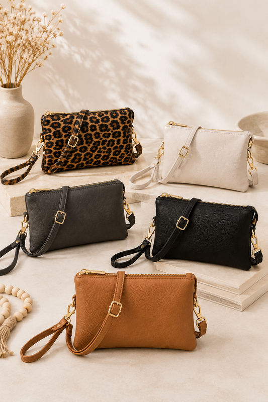 Five mini shoulder bags in various colors and textures are arranged on a beige surface with decorative props including a vase of dried flowers, books, and a beaded bracelet. The bags include leopard print, off-white, dark gray, black, and tan/cognac, each with a wristlet strap and gold-tone hardware.