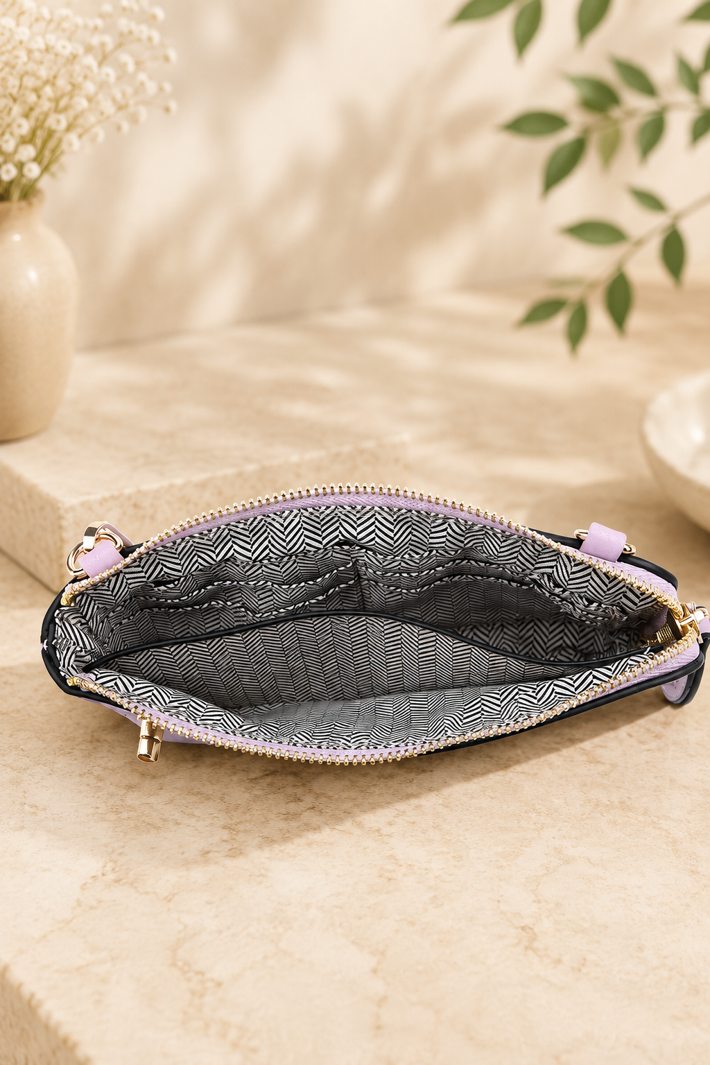 An open zippered pouch with a black and white geometric patterned interior lining and light purple zipper pulls, displayed on a beige stone surface with soft-focus decorative background elements.