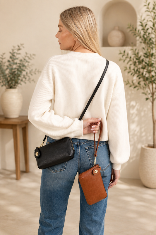 A woman shown from the back wearing a cream ribbed knit sweater, blue jeans, and carrying a black crossbody bag and a brown pouch with a phone strap in a warm, neutral-toned interior setting.