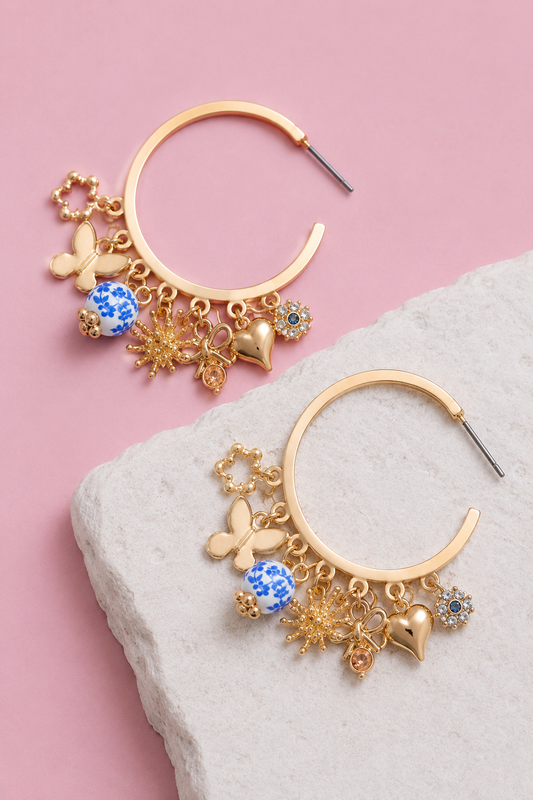 Two gold hoop earrings with blue floral porcelain beads and metal charms on a white stone and pink background.