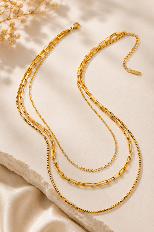 Three layered gold metal chains in box, link, and cable styles displayed on a white stone surface.