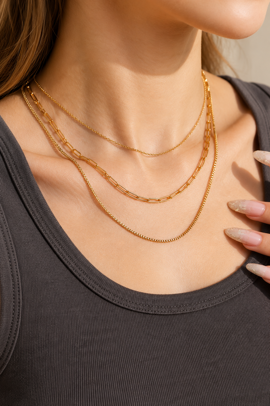 Three layered gold metal necklaces including a paperclip chain and two thin link chains worn on a person.