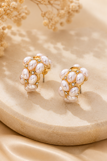 A pair of gold-toned cluster earrings decorated with white baroque pearls and small clear rhinestones, displayed on a beige stone slab with dried flowers in the background.