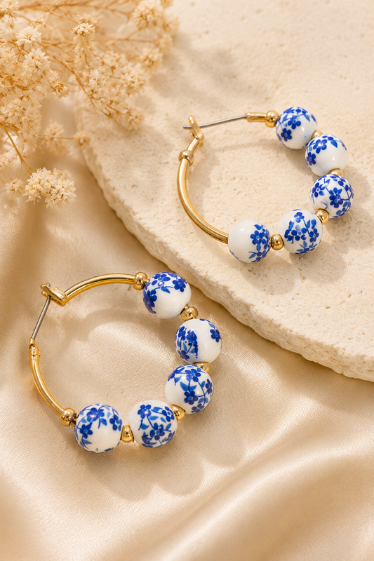 Gold hoop earrings with white ceramic beads featuring a blue floral pattern, styled on a beige satin fabric with dried flowers.