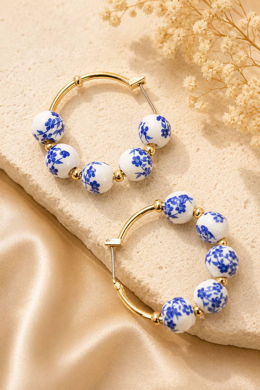 Gold hoop earrings with white ceramic beads featuring a blue floral pattern, displayed on a beige stone slab with dried flowers and satin fabric.