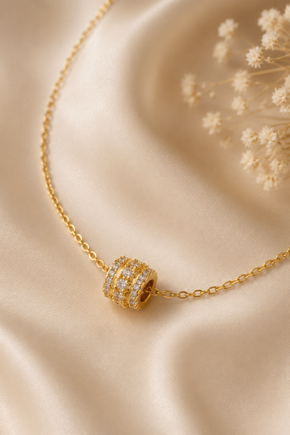 Gold metal chain necklace with a barrel-shaped slider pendant featuring three rows of clear stones on draped fabric.
