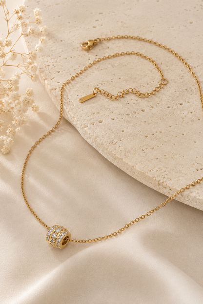 Gold chain necklace with a barrel-shaped pendant featuring three rows of clear stones on a stone and fabric background.