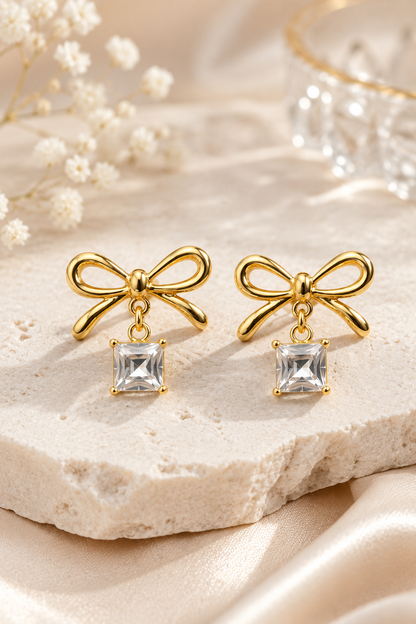 Pair of gold bow earrings with square crystal drops resting on a white stone surface.
