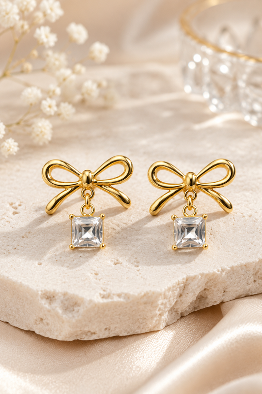 Pair of gold bow earrings with square crystal drops resting on a white stone surface.