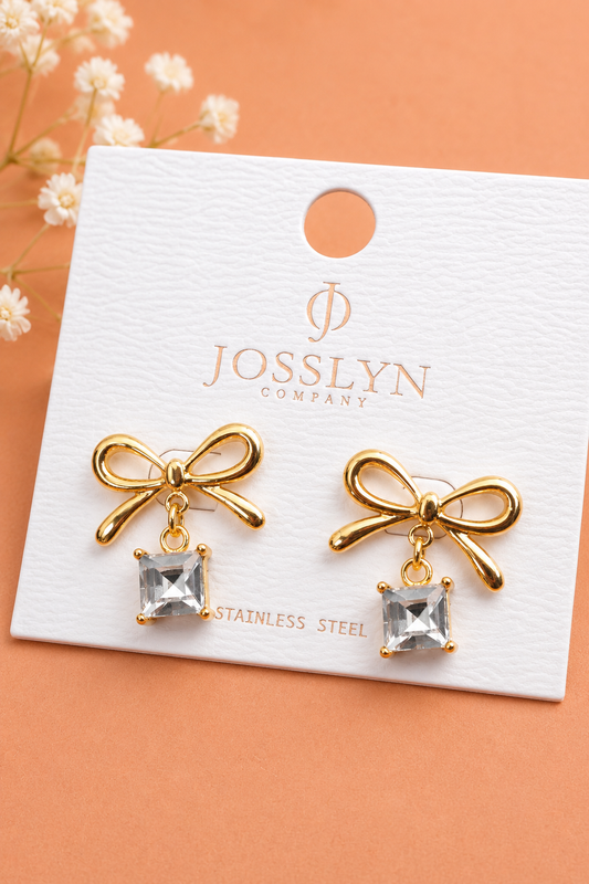 Two gold metal bow earrings with square crystal drops on a white Josslyn COMPANY card printed with STAINLESS STEEL.