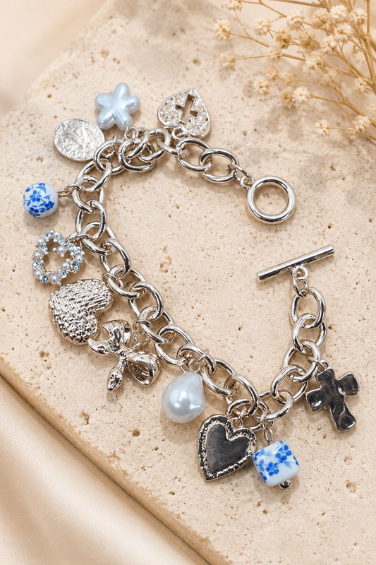 Gold charm toggle bracelet with blue porcelain hearts, cross, pearl, bow, floral, and crystal charms. Flat Lay