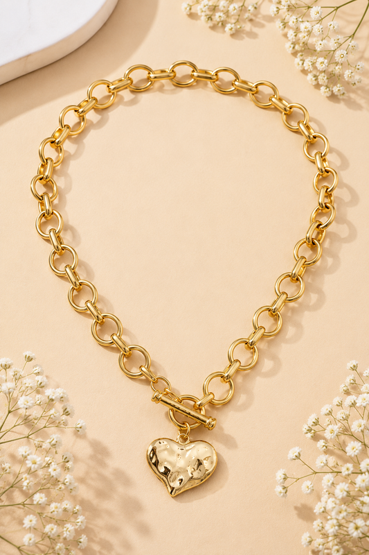 A gold chain necklace with a toggle clasp and a dangling heart-shaped charm, styled on a warm beige background with small white flowers.