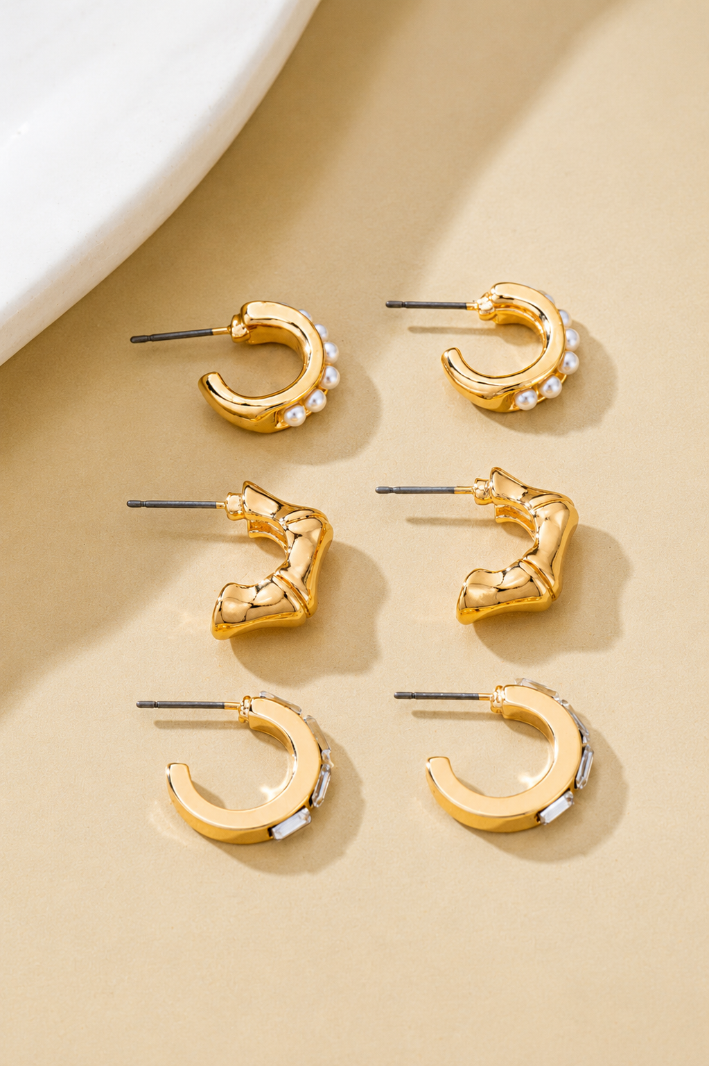 Gold hoop earring trio set featuring pearl huggies, sculptural C-hoops, and baguette crystal hoops. Flat lay 2