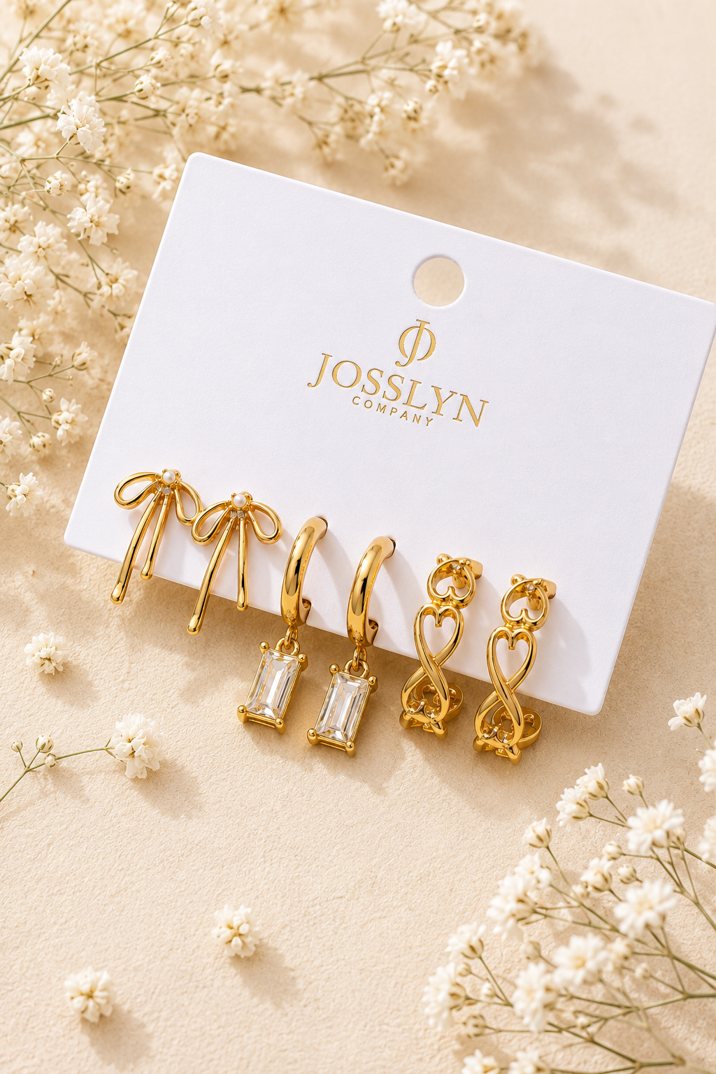 A set of five gold-toned hoop earrings displayed on a white Josslyn Company card, surrounded by small white flowers on a warm beige surface.