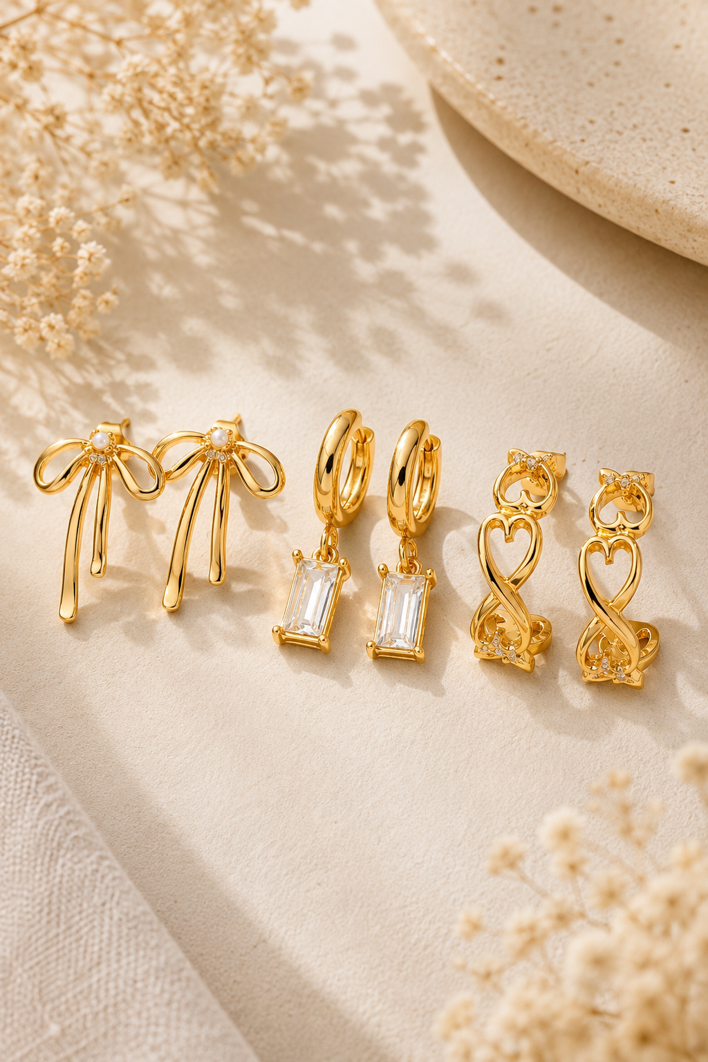 A set of gold-toned jewelry pieces arranged on a light beige fabric surface with dried floral props. The set includes bow-shaped earrings, hoop earrings with rectangular clear stones, heart-shaped earrings, and a small infinity symbol earring.