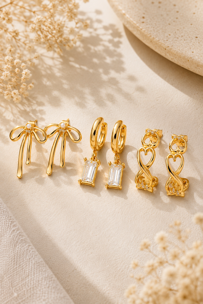 A set of gold-toned jewelry pieces arranged on a light beige fabric surface with dried floral props. The set includes bow-shaped earrings, hoop earrings with rectangular clear stones, heart-shaped earrings, and a small infinity symbol earring.