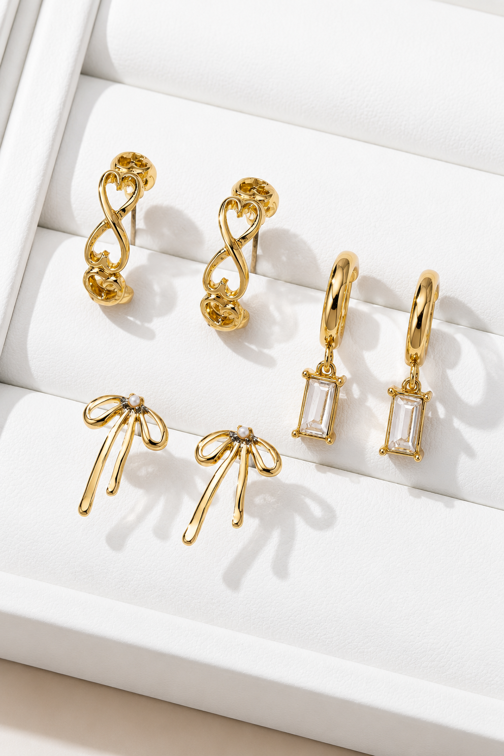A set of six gold-toned earrings displayed on a white jewelry box. The collection includes two twisted infinity hoop earrings, two dangling earrings with rectangular clear stones, and two bow-shaped earrings with small clear stones, all arranged on a white padded tray.